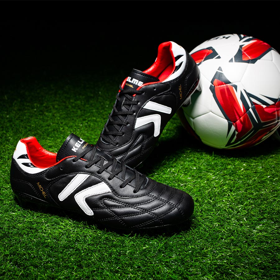 Kelme Black soccer cleats with red and white accents on a grass field with a soccer ball.