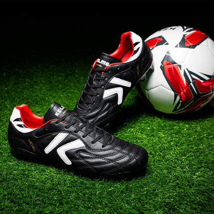 Kelme Black soccer cleats with red and white accents on a grass field with a soccer ball.